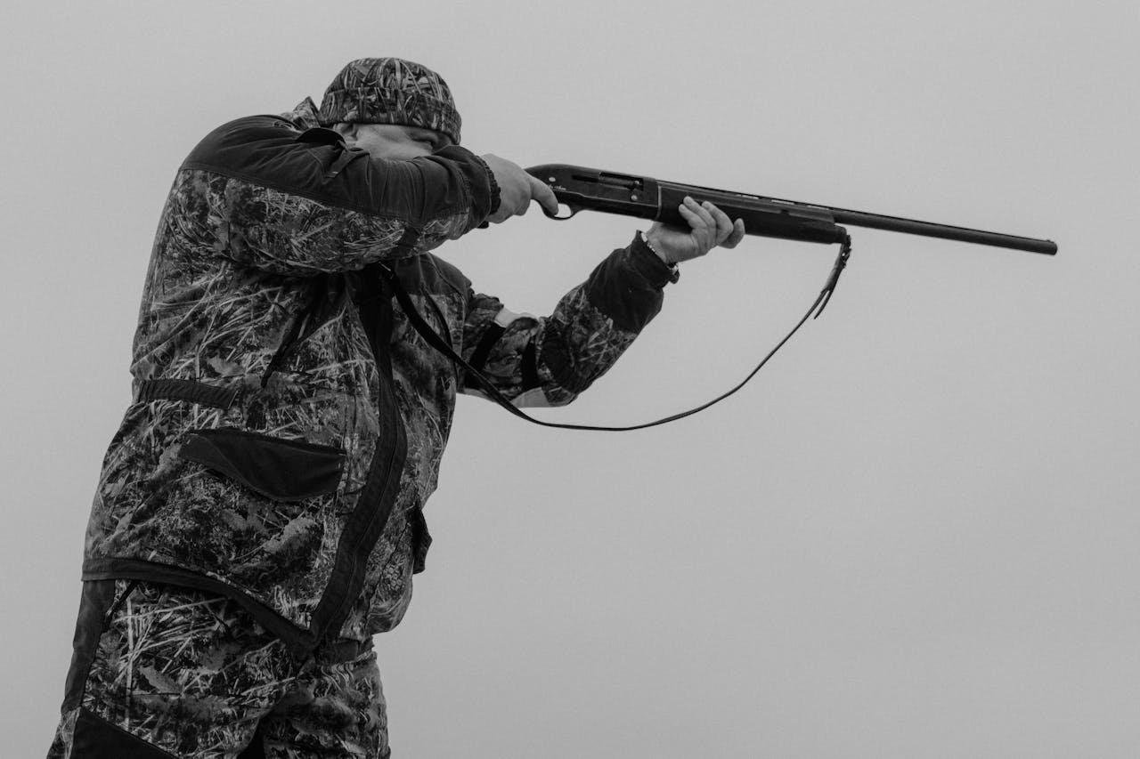 Black and white image of a hunter aiming a rifle, dressed in camouflage, against a cloudy sky.